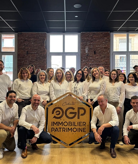 Équipe Cabinet OGP Immobilier Patrimoine posant souriante autour de leur logo hexagonal en bois. Locaux modernes, murs briques.