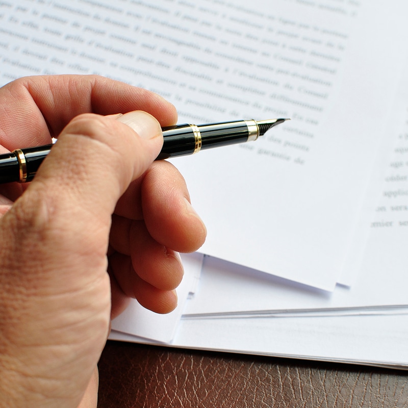 Main avec stylo plume noir et or sur documents texte, évoquant évaluation ou décision importante.