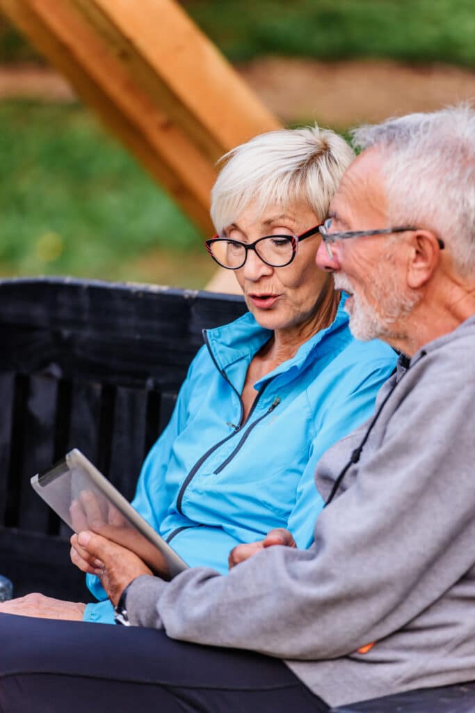 Couple âgé souriant et engagé, assis dehors, regardant ensemble une tablette numérique. La femme porte des lunettes originales.