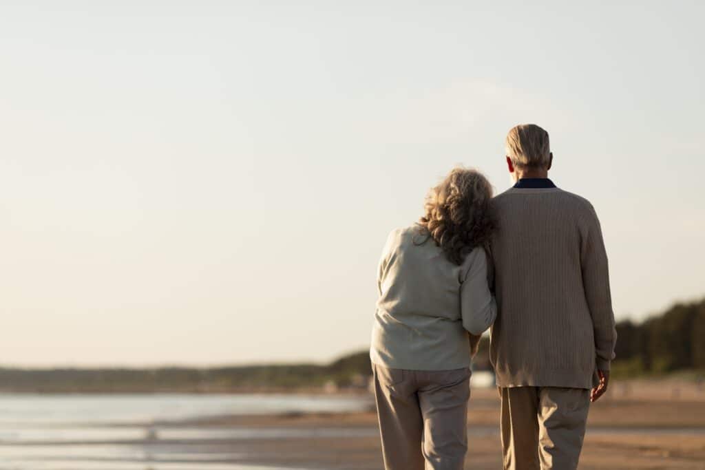 Couple âgé de dos marchant sur une plage au coucher du soleil. La femme s'appuie tendrement sur l'épaule de l'homme.