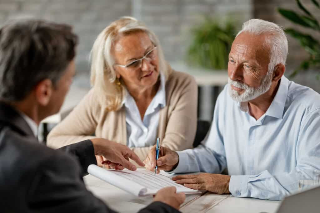 Un couple senior signe un contrat important avec un conseiller financier. L'homme écrit tandis que la femme aux lunettes regarde attentivement.