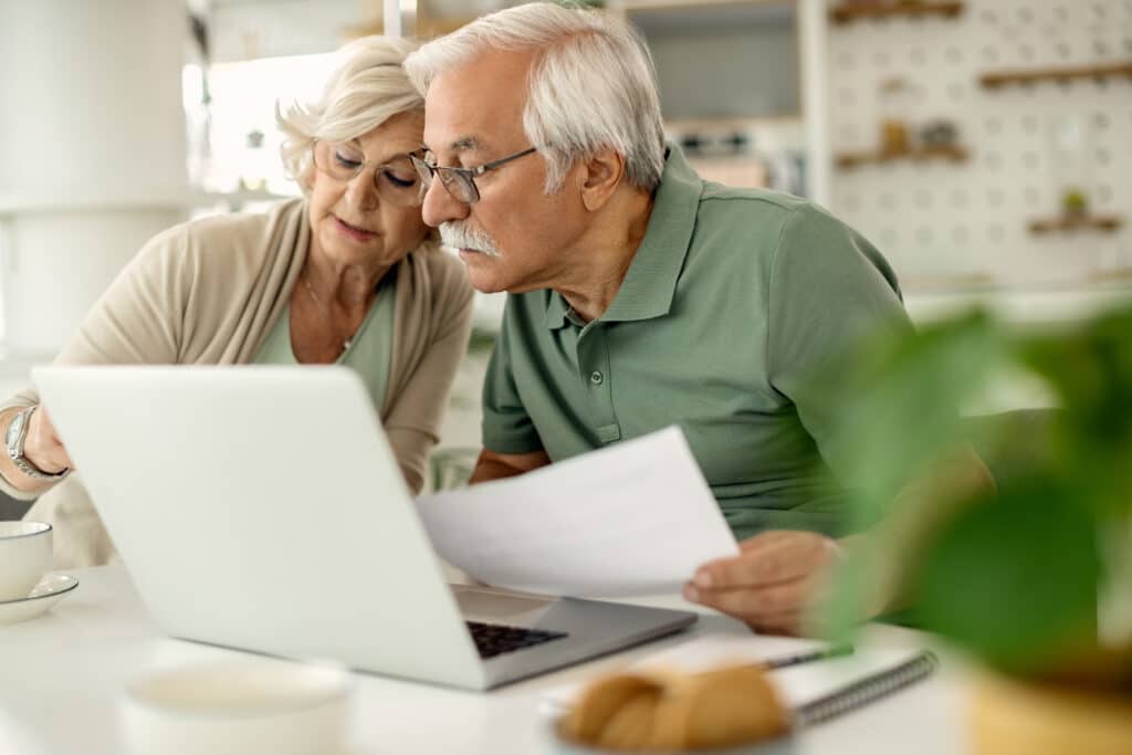 Couple âgé aux cheveux blancs, lunettes, examine un ordinateur portable et des documents. Ils gèrent leurs finances à domicile.